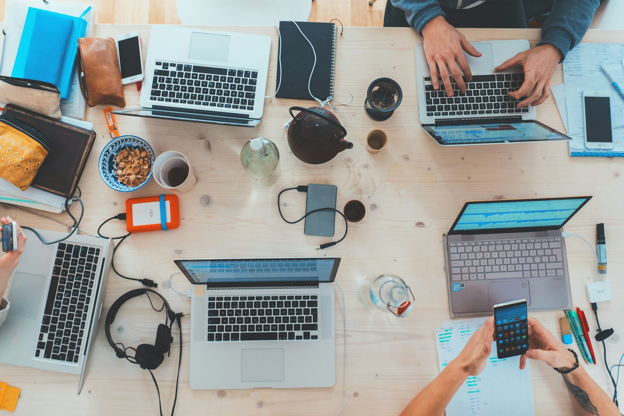Overhead view of a shared work table covered with laptops, notebooks, and tea, hands typing at the keyboards